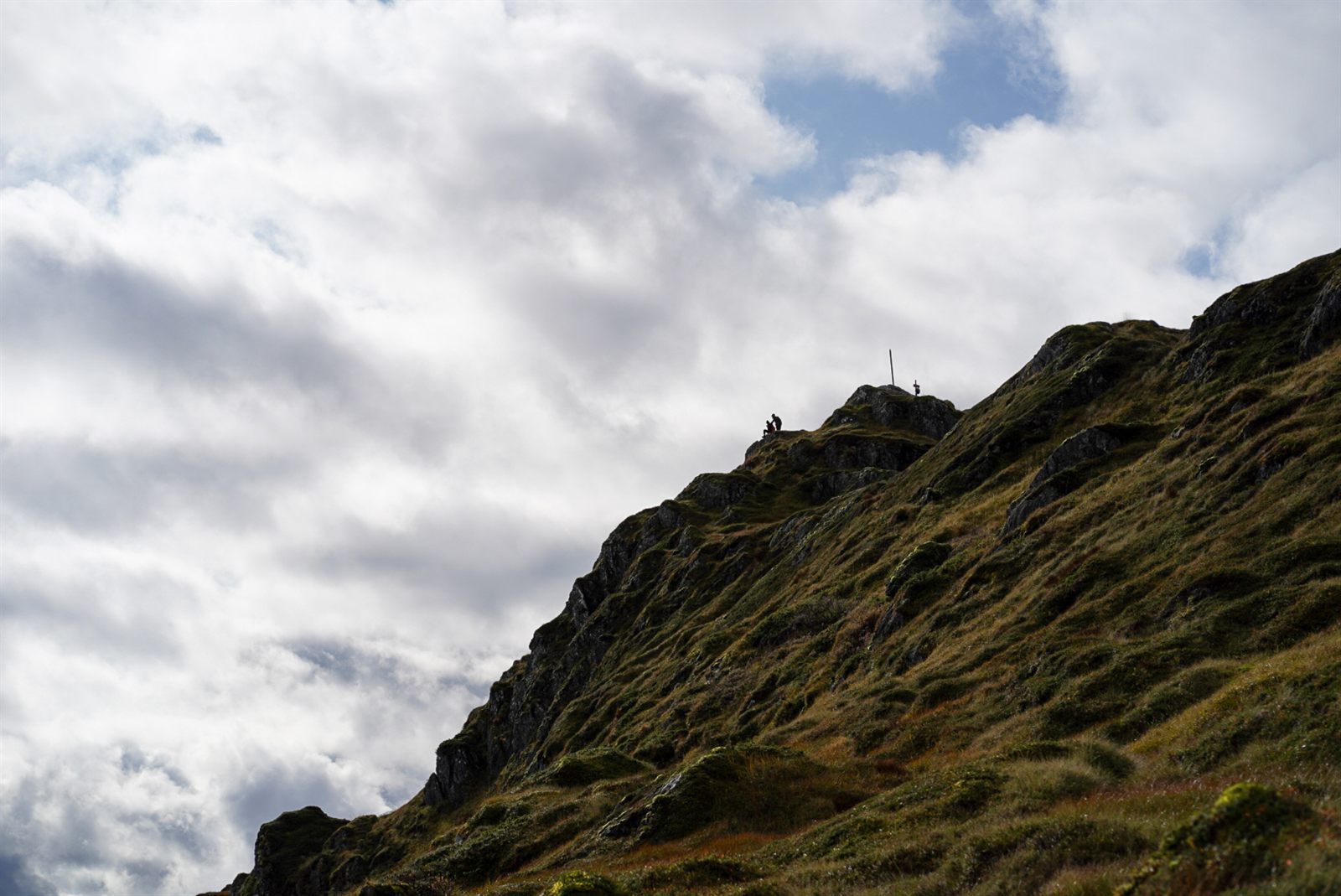 Two tiny hikers silhouetted at the summit of a rocky mountain ridge against a cloudy sky